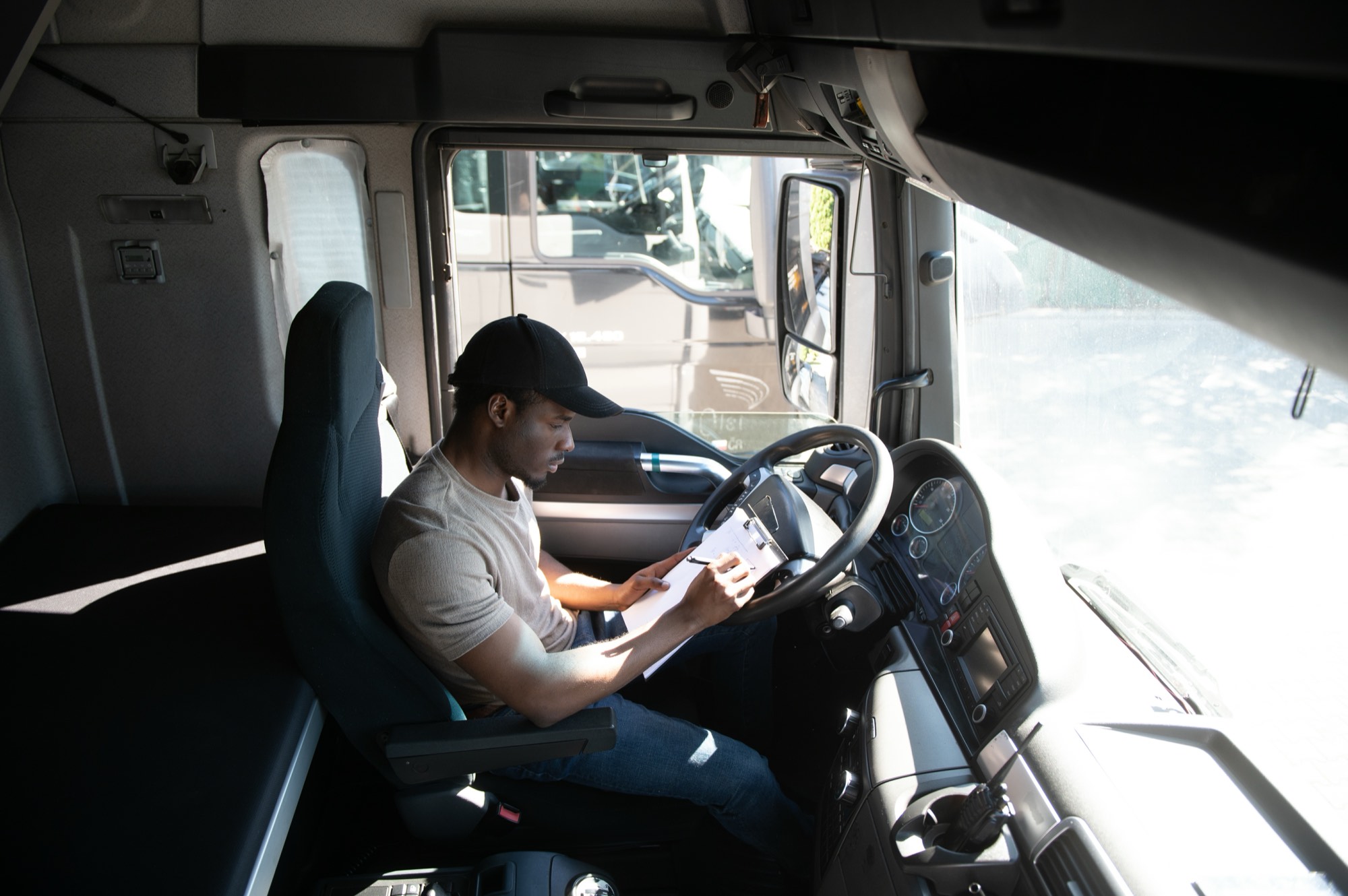 A truck driver sitting in a parked cab filling out paperwork while waiting