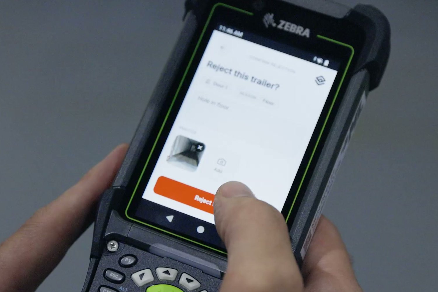 A rugged handheld terminal held in two hands, asking whether to reject this trailer, with a photo attached and an orange reject button